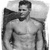 Static grayscale image of a shirtless man with short dark hair and facial stubble, standing in profile facing left against a plain background.