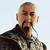 Pixelated image of a person with a shaved head wearing a dark high-collared jacket over a light patterned shirt against a light background.