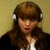 A person with brown hair and bangs wears white headphones, a black jacket over a white shirt, against a beige background.