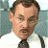 Pixelated close-up of a person with short brown hair wearing a white shirt and dark tie against a light blue background.