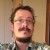 Pixelated static image of a person with short dark hair, light skin, and a mustache, wearing a checkered shirt against a neutral background.