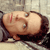 A pixelated image of a person lying on their back with eyes closed, mouth open, and a red mark on their forehead against a light background.