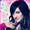 A woman with dark hair and blue eyes wears a pink top, her hand near her chest, with a pink heart symbol on a light background.