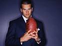 Static image of a man in formal attire holding an American football against a dark blue background.