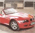 A red convertible car with a white soft top is positioned at an angle, viewed from the front-left side, against a blurred background of indistinct shapes.