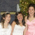Three women in formal dresses stand outdoors; one wears a pink dress, two wear white dresses, with green foliage in the background.