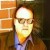 Pixelated static image of a person with light skin tone, wearing glasses and a dark jacket over a light shirt, positioned against a blurred background with warm orange and brown tones.