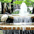 Pixelated image of a white fountain with water jets, surrounded by green foliage and dark tree trunks, with a reflective water surface below.