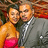 Two people in close proximity, one in orange top and pink bottom, the other in gray suit with white shirt and dark tie, against a blurred background with warm tones.