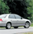 A silver sedan with black wheels and a red taillight is positioned on a road, surrounded by green foliage and dark trees in the background.
