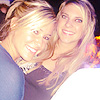 Two women with blonde hair pose closely together, both smiling broadly with visible teeth, one wearing a dark top with a silver necklace, the other in a light-colored top.