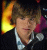 Static pixelated image of a person with brown hair wearing a dark jacket over a light shirt, set against a black background with yellow and orange gradient on the left side.