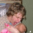 A child with brown hair in a pink dress and blue collar, holding hands near the chest against a blurred background.