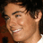 Pixelated close-up of a smiling person with short dark hair, visible ear, and light skin tone against a dark background.
