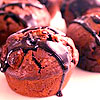 A close-up of a chocolate muffin with dark chocolate drizzle, surrounded by similar muffins in a blurred background.