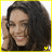 A pixelated image of a person with long dark hair, wearing a black top and yellow border, smiling while holding their hand near the head.