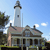 Pixelated static image of a white lighthouse with dark trim atop a multi-story building under a blue sky with scattered clouds.