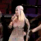 Animated sequence of a person with long blonde hair in a white outfit performing on stage with a microphone, surrounded by backup dancers in dark brown outfits.