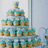 A multi-tiered cupcake tower with turquoise frosting and yellow bases on silver stands, displayed against a light blue background.