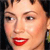 A close-up of a person's face with short dark hair, red lips, and a slight smile, set against a dark background.