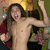 A shirtless person with long brown hair raises both arms in a celebratory pose against a blurred background of green, red, and yellow tones.