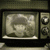 A static image of a vintage television set with a grainy screen displaying an indistinct figure, framed by dark borders and control knobs.