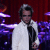 Pixelated static image of a person in white shirt and black tie against red background with purple drum set elements visible.