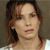 Pixelated close-up of a person with light brown hair, fair skin, and a neutral expression wearing a white top against a muted brown background.