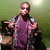 Pixelated image of a person in purple sleeveless top and dark jacket holding drumsticks near drums against green background.