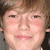 Pixelated close-up of a person's face with light brown hair, pink cheeks, and closed mouth showing teeth.