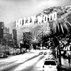 Black-and-white photograph of a street scene with the Hollywood sign visible on a hillside, cars on the road, and buildings in the background.