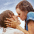 Two people kiss under a blue sky with clouds, one wearing a white top and the other in a blue sleeveless shirt.