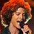 Pixelated static image of a person with voluminous curly red hair singing into a microphone, wearing dark clothing against a dark background.