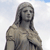 Pixelated grayscale image of a figure with long hair, wearing a draped garment, set against a light background with faint blue sky at top.