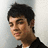 Pixelated static image of a person with short dark hair wearing a dark jacket over a light shirt against a light gray background.
