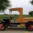 Pixelated image of a vintage open-top vehicle with brown body and dark wheels on grassy terrain under overcast sky.