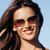 Pixelated close-up of a person with long brown hair, wearing dark sunglasses, smiling against a light blue background.