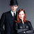 Two figures in formal attire stand side-by-side against a gray background; the taller figure wears a black hat and dark suit with a white shirt, while the shorter figure has long brown hair and a dark top.