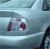 A pixelated image of a car's side with a visible headlight and part of the front fender, featuring a small dark red or maroon emblem on the vehicle's body.