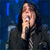 A person with long dark hair sings into a microphone against a blue background, next to a static portrait of the same individual in front of a light backdrop.