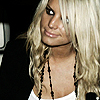 A static image of a person with long blonde hair wearing a black choker necklace and a white top, with a dark background.