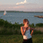 Pixelated static image of a person standing outdoors near water, wearing a sleeveless shirt and dark pants, with a sailboat on the water and a cloudy sky in the background.