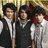 Three individuals in dark vests and white shirts stand together against a blurred background with colorful heart shapes scattered across the image.