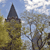 Pixelated image of a tall dark spire atop a building with yellowish-brown walls, surrounded by trees under a blue sky with white clouds.
