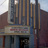 Pixelated image of a theater facade with vertical columns, a marquee displaying red text, and a dark entrance below.
