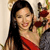A woman with long dark hair wears a red dress and holds a bouquet of red flowers while smiling at the camera.