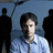 Pixelated static image of a person in a light blue collared shirt against a blue background with dark silhouettes on both sides.