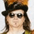 Pixelated portrait of a person wearing dark sunglasses, a black cap, and long brown hair against a light background with orange-brown tones at the top.