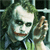 Pixelated image of a character with white face paint, dark eye makeup, and a red smile, holding a small object near the face against a green background.