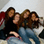 Four young women sit closely together on a light-colored couch with red accents, wearing dark tops and blue jeans, in a pixelated image with visible motion blur.